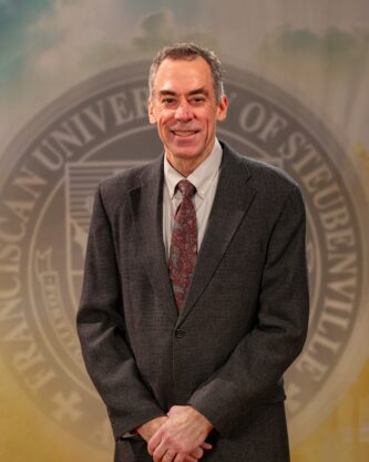 Dr. Ron Bolster_Faculty Portrait_Spring2026_-2816 Portrait of Dr. Ron Bolster standing in front of the Franciscan University of Steubenville seal.