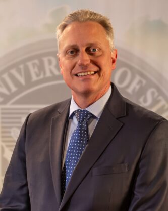 To Web - Dr. John Bergsma FEB2025 --4 Dr. John Bergsma standing in front of the Franciscan University of Steubenville seal.