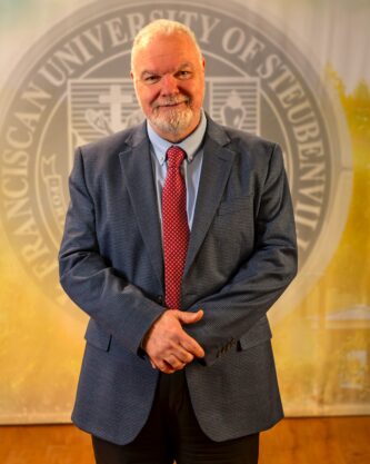 Dr. Petroc Willey standing in front of a university seal.