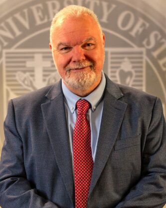 Dr. Petroc Willey standing in front of a university seal.