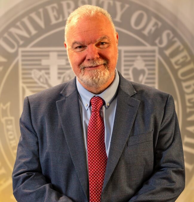 To Web – Dr. Petroc Willey FEB2025 –6 Dr. Petroc Willey standing in front of a university seal.